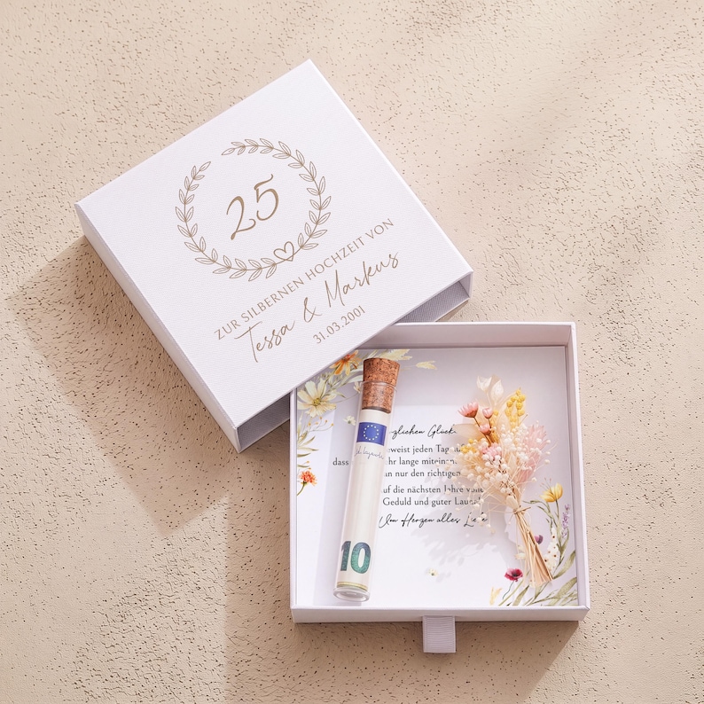 Pu&ograve; includere: Una scatola regalo quadrata bianca con coperchio scorrevole decorata con una corona d'alloro dorata, il numero 25 e il testo "ZUR SILBERNEN HOCHZEIT VON Tessa & Markus 31.03.2001". All'interno, una fiala di vetro con tappo in sughero e banconota.