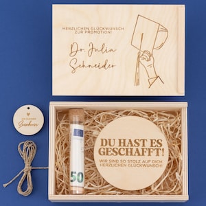 Könnte beinhalten: Eine hölzerne Geschenkbox mit Deckel, die ein rundes Holzstück mit Text, ein Glasröhrchen mit einem 50-Euro-Schein und dekoratives Stroh enthält. Der Deckel hat Text und eine Illustration einer Abschlusskappe. Ein kleines Holzschild und Bindfaden sind ebenfalls enthalten.