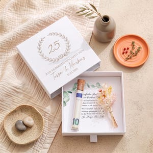 Pu&ograve; includere: Una scatola regalo quadrata bianca con cassetto scorrevole, decorata con una corona d'alloro dorata, il numero 25 e il testo "ZUR SILBERNEN HOCHZEIT VON Tessa & Markus 31.03.2001". All'interno, una fiala di vetro con tappo, una banconota e fiori secchi.