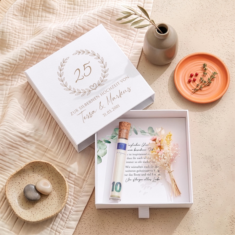 Pu&ograve; includere: Una scatola regalo quadrata bianca con cassetto scorrevole, decorata con una corona d'alloro dorata, il numero 25 e il testo "ZUR SILBERNEN HOCHZEIT VON Tessa & Markus 31.03.2001". All'interno, una fiala di vetro con tappo, una banconota e fiori secchi.