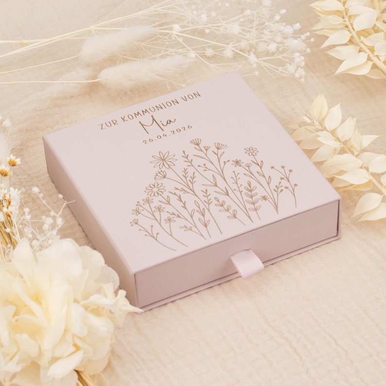 May include: A light pink gift box with a floral design and the name "Mia" printed on the lid. The box has a ribbon pull and text that reads "ZUR KOMMUNION VON" and the date "26.04.2026".