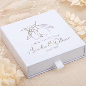 May include: A square white sliding gift box featuring a gold-colored line art illustration of two lemons on a branch. The text reads ZUR HOCHZEIT VON Annika & Oliver 08.08.2026 in a mix of serif and script fonts. A small white fabric pull tab is attached.