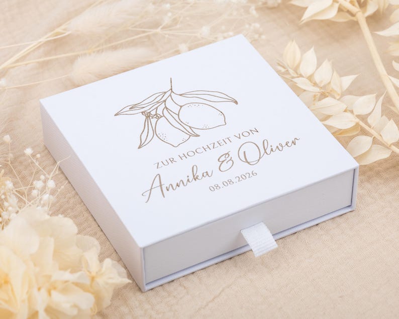 May include: A square white sliding gift box featuring a gold-colored line art illustration of two lemons on a branch. The text reads ZUR HOCHZEIT VON Annika & Oliver 08.08.2026 in a mix of serif and script fonts. A small white fabric pull tab is attached.