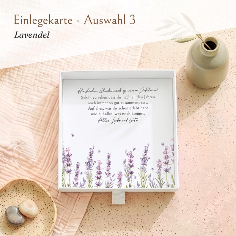 Pu&ograve; includere: Una scatola regalo quadrata bianca con un coperchio che presenta un'illustrazione floreale di lavanda sul fondo e il testo in tedesco: Herzlichen Gl&uuml;ckwunsch zu eurem Jubil&auml;um! Sch&ouml;n zu sehen, dass ihr nach all den Jahren noch immer so gut zusammenpasst.