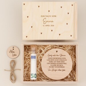 May include: A wooden keepsake box with a lid featuring small heart designs and the text "ZUR TAUFE VON Emma 12. April 2026". Inside, a round wooden piece with engraved text and a glass tube with a "50" label.
