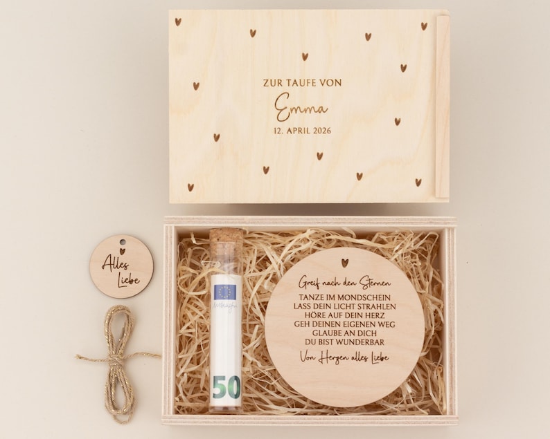 May include: A wooden keepsake box with a lid featuring small heart designs and the text "ZUR TAUFE VON Emma 12. April 2026". Inside, a round wooden piece with engraved text and a glass tube with a "50" label.