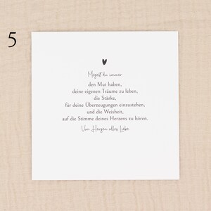 May include: A white card with black text and a small heart illustration. The card features a handwritten font with a message in German. The number "5" is in the upper left corner. The card is on a beige fabric background.