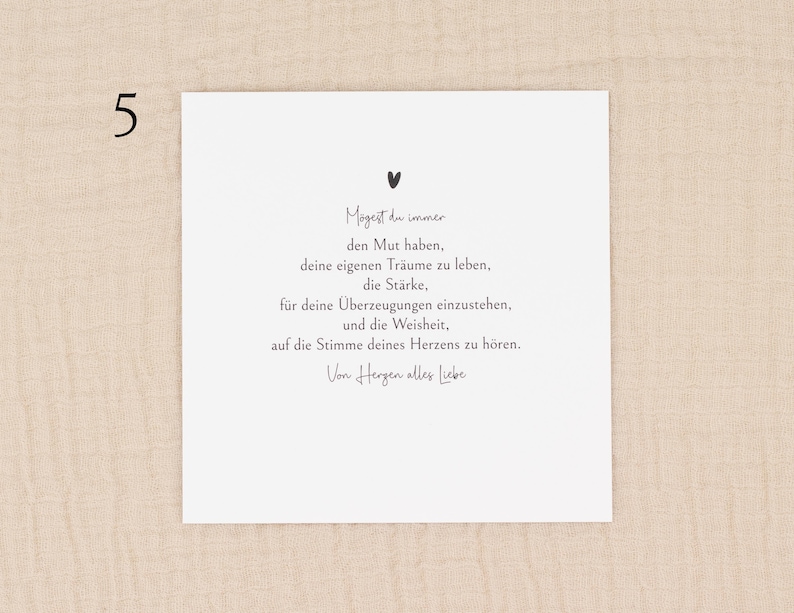 May include: A white card with black text and a small heart illustration. The card features a handwritten font with a message in German. The number "5" is in the upper left corner. The card is on a beige fabric background.