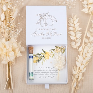May include: A white rectangular gift box with a sliding drawer containing a glass tube with a cork stopper holding a ten euro banknote, a card with lemon illustrations and German text, and a small dried floral sprig, with dried flowers nearby.