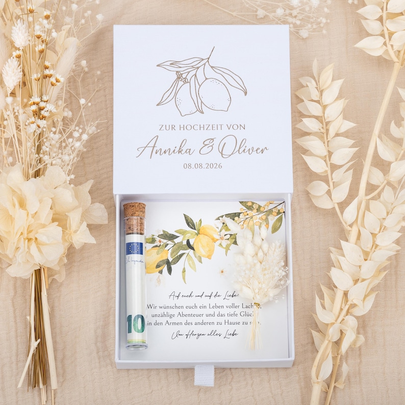 May include: A white rectangular gift box with a sliding drawer containing a glass tube with a cork stopper holding a ten euro banknote, a card with lemon illustrations and German text, and a small dried floral sprig, with dried flowers nearby.
