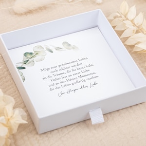 May include: A square white gift box with a sliding drawer featuring a white card insert. The card displays a watercolor eucalyptus branch illustration and German text that reads: M&ouml;ge euer gemeinsames Leben noch sch&ouml;ner werden als die Tr&auml;ume, die ihr heute habt. Haltet fest an eurer Liebe und an den kleinen Momenten, die das Leben gro&szlig;artig machen. Von Herzen alles Liebe.