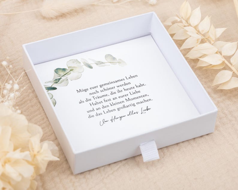 May include: A square white gift box with a sliding drawer featuring a white card insert. The card displays a watercolor eucalyptus branch illustration and German text that reads: M&ouml;ge euer gemeinsames Leben noch sch&ouml;ner werden als die Tr&auml;ume, die ihr heute habt. Haltet fest an eurer Liebe und an den kleinen Momenten, die das Leben gro&szlig;artig machen. Von Herzen alles Liebe.