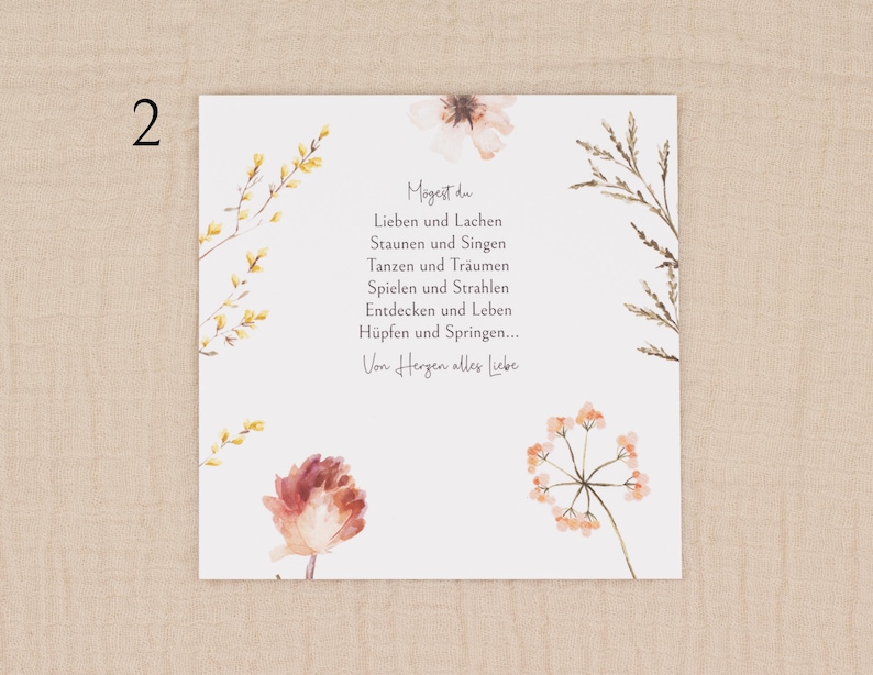 May include: A square card with watercolor floral illustrations and text in German. The card features a white background with delicate flowers in shades of peach, yellow, and brown. The text is centered and reads "Mogest du..." followed by a poem.