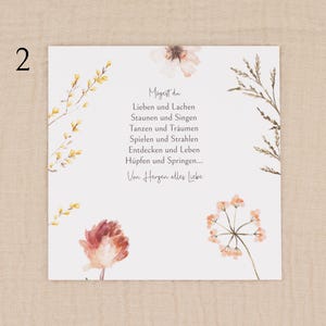 May include: A square card with watercolour floral illustrations and German text. The card has a white background with delicate flowers in peach, yellow, and brown. The text is centred and reads "Mogest du..." followed by a poem.