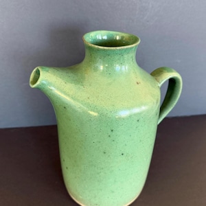 May include: A ceramic pitcher with a speckled light green glaze. The vessel features a cylindrical body, a short neck with a wide opening, a small pouring spout on the side, and a curved handle attached to the opposite side of the spout.