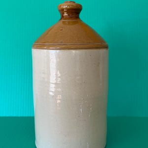 Pode incluir: Uma jarra cilíndrica de grés com um corpo de cor creme brilhante e um ombro e gargalo esmaltados em castanho contrastante. O recipiente tem uma abertura estreita no topo, um acabamento liso e uma base resistente com um design funcional.