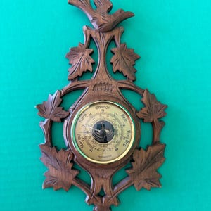 May include: A wooden barometer with a bird and leaf design. The barometer face is round with a gold-colored rim and features a needle pointing to the current weather conditions. The text on the barometer face includes "Rain", "Change", "Fair", and "Very Dry".