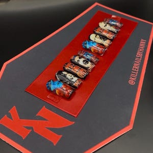 Peut inclure: Un ensemble de dix faux ongles en forme de cercueil avec divers motifs, notamment de l'art abstrait et des motifs de crânes. Les ongles sont disposés sur une carte rectangulaire rouge, placée sur un fond noir avec le texte "KN" en rouge. Le compte Instagram @KILLERNAILSBYHANNY est visible.