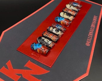 May include: A set of ten coffin-shaped false nails with various designs, including abstract art and skull motifs. The nails are arranged on a red rectangular card, placed on a black background with the text "KN" in red. The Instagram handle @KILLERNAILSBYHANNY is visible.