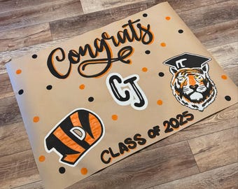 May include: A brown paper graduation sign with the word "Congrats" in black and orange script. The sign includes the initials "CJ", a tiger wearing a graduation cap, and the words "Class of 2025". Orange and black polka dots are scattered across the sign.