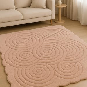 Puede incluir: Una alfombra de color rosa pálido con un diseño de espirales. La alfombra tiene un aspecto suave y texturizado y un borde festoneado. Está colocada sobre un suelo de madera en un salón, cerca de un sofá beige.