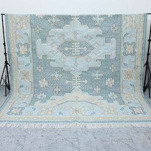 Puede incluir: Una alfombra rectangular grande con un diseño geométrico central en tonos azul claro y crema. La alfombra presenta un patrón detallado con motivos florales y geométricos, bordeada por un borde color crema y terminada con una franja. La alfombra se muestra sobre un fondo blanco.