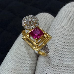 May include: A gold ring with a square, magenta gemstone in a diamond-shaped frame. Above the magenta stone is a cluster of small, clear stones. The band features more small stones. The ring is a piece of jewellery.