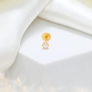 May include: A single gold earring with a small, round, white pearl-like center. The earring has a post and a small, decorative design around the pearl. The earring is set against a white background.