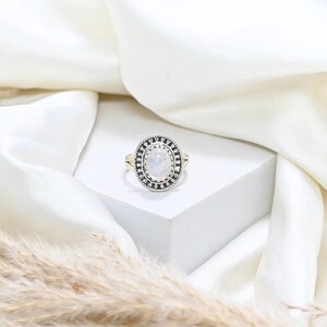 May include: A silver and gold ring featuring an oval, iridescent gemstone. The gemstone is framed by a black and silver border with a decorative pattern. The ring is displayed on a white surface.