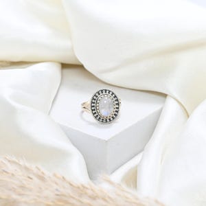 May include: A silver ring featuring an oval, iridescent gemstone. The ring has a detailed, textured border and a split shank. The ring is presented on a white surface with a white fabric backdrop.