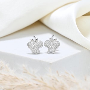 May include: A pair of silver-toned apple-shaped stud earrings. Each earring is encrusted with small, sparkling stones, creating a shimmering effect. The earrings have a detailed design, with a stem and leaf detail at the top. The earrings are displayed on a white surface.