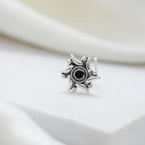 Puede incluir: Un piercing de nariz de plata con una piedra negra redonda central engastada. El metal tiene un acabado oxidado y un diseño en forma de estrella compuesto por rayos curvos y puntiagudos con pequeños acentos esféricos en la base.