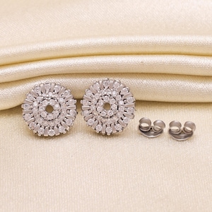 May include: Two silver-coloured, round stud earrings with a circular design of small, clear stones. The earrings are next to two earring backs. The earrings are on a cream-coloured, textured surface.