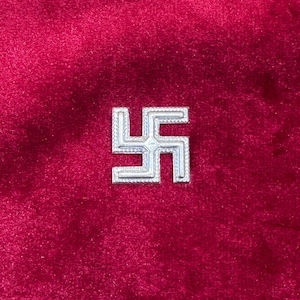 Könnte beinhalten: Ein silbernes Hakenkreuz-Emblem, zentriert auf einem tiefroten Samthintergrund. Das Design des Emblems besteht aus vier rechtwinklig gebogenen Armen, die eine symmetrische kreuzartige Form bilden. Der Samt hat ein weiches, strukturiertes Aussehen.