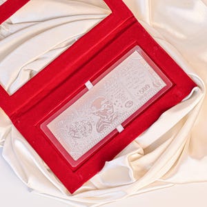 Può includere: Una barra d'argento che assomiglia a una banconota da 500 rupie, esposta in una custodia di velluto rosso. La barra presenta un'immagine incisa di Mahatma Gandhi e dettagli intricati. La custodia è posta su uno sfondo di tessuto bianco.