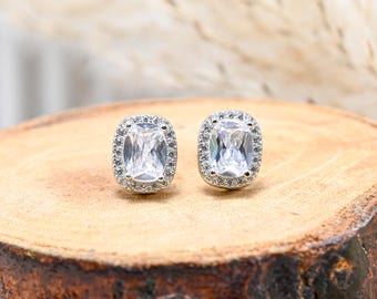 May include: A pair of silver-toned stud earrings. Each earring features a large, rectangular, clear gemstone surrounded by a halo of smaller, clear stones. The earrings are displayed on a wooden surface.