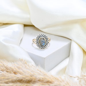 May include: A gold-toned ring featuring an oval-shaped, iridescent gemstone center, surrounded by a halo of small, silver-colored beads. The ring's band has a split design. The ring is displayed on a white surface with a soft, white fabric backdrop.