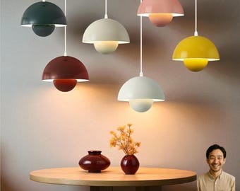 May include: A selection of contemporary pendant lights in shades of green, white, red, pink, and yellow. Each light features a rounded shade and a spherical base, hanging from white cords. The lights are positioned above a wooden table with vases.
