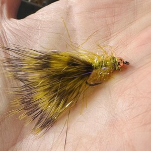May include: A handmade fly fishing lure with a hook. The lure features a combination of yellow, brown, and black feathers, with a touch of iridescent material. The head is a small, orange bead. This fly is designed for catching fish.