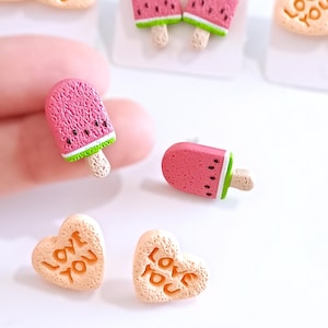 Pode incluir: Close-up de pingentes em miniatura de picolés de melancia e formatos de coração. Os picolés são rosa com sementes pretas e borda verde, em um palito de madeira. Os pingentes de coração são bege com as palavras "LOVE YOU" em laranja.