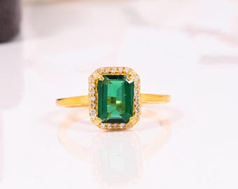 May include: A gold ring with a rectangular, emerald-cut green gemstone. The gemstone is framed by a halo of small, clear stones. The ring has a simple, elegant band.