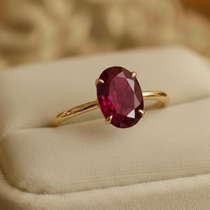 Può includere: Un anello d'oro con una gemma ovale rosso intenso. L'anello è posto su un portagioie bianco foderato di velluto. Lo sfondo è un morbido mix sfocato di beige e oro, che mette in risalto l'elegante design dell'anello.