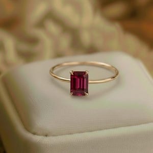 May include: A gold ring with a rectangular, deep red gemstone. The ring is displayed on a white surface, with a soft, blurred background. The gemstone is the focal point, showcasing its rich colour and cut.