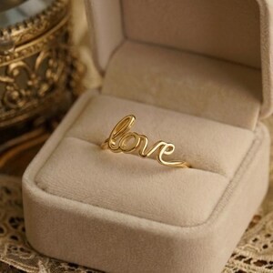 Può includere: Un anello color oro con la parola "love" in corsivo. L'anello è esposto all'interno di un portagioie beige con un interno morbido e vellutato. L'anello è un gioiello delicato.