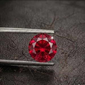May include: A round, brilliant-cut vibrant red gemstone held by a pair of silver-toned metal tweezers. The gemstone features a faceted surface that reflects light, creating a bright white sparkle on its upper edge against a dark textured surface.