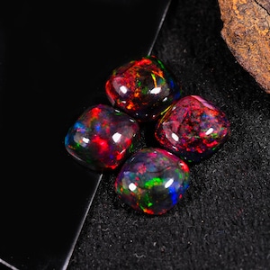 May include: Four polished black opal gemstones displaying flashes of red, green, and blue. The opals are rounded squares with a glossy finish, set against a dark background.