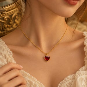 May include: A gold chain necklace featuring a single heart-shaped pendant with a faceted deep red gemstone. The pendant is set in a gold frame and hangs from a delicate gold link chain, resting against the collarbone area of the wearer.