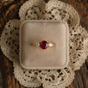 Peut inclure: Une bague en or ornée d'une grande pierre rouge ronde à facettes au centre, flanquée de deux pierres blanches rondes et transparentes de chaque côté. La bague est montée sur un anneau en or poli et lisse, présentant un design à trois pierres.