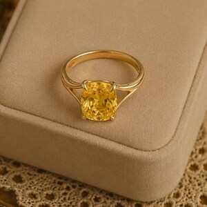 May include: A gold ring featuring a cushion-cut yellow gemstone set in a four-prong mounting. The band has a split shank design that tapers towards the centre stone. The ring is displayed on a smooth, light beige surface.