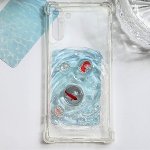 May include: Clear phone case with a blue resin design featuring two red fish, bubbles, and seashells. The case has a clear border and a rectangular design with a wave-like pattern. The design is reminiscent of an underwater scene.
